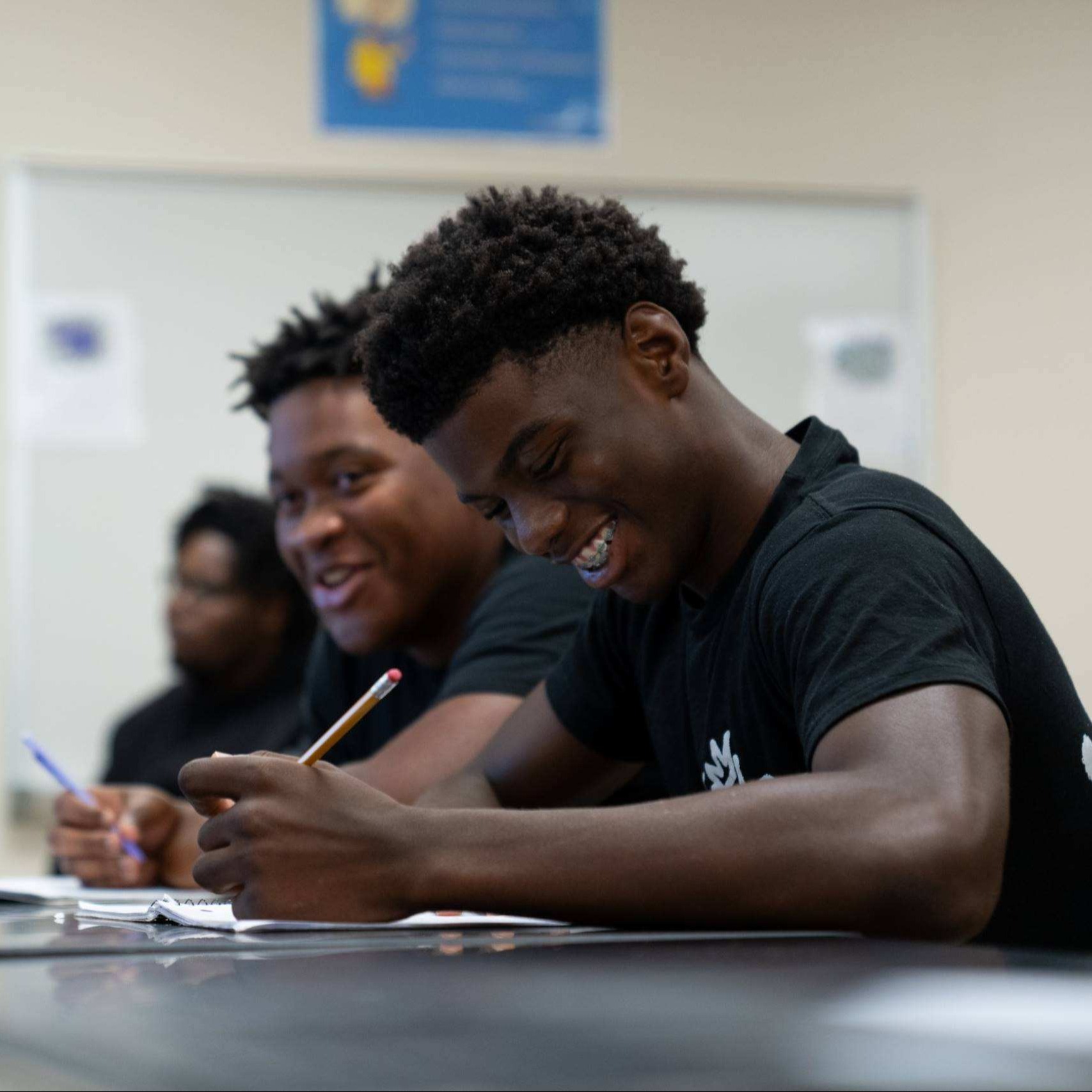 Two men in black t-shirts sit at a table in a classroom, writing in notebooks. Behind them, a whiteboard is mounted on the wall.