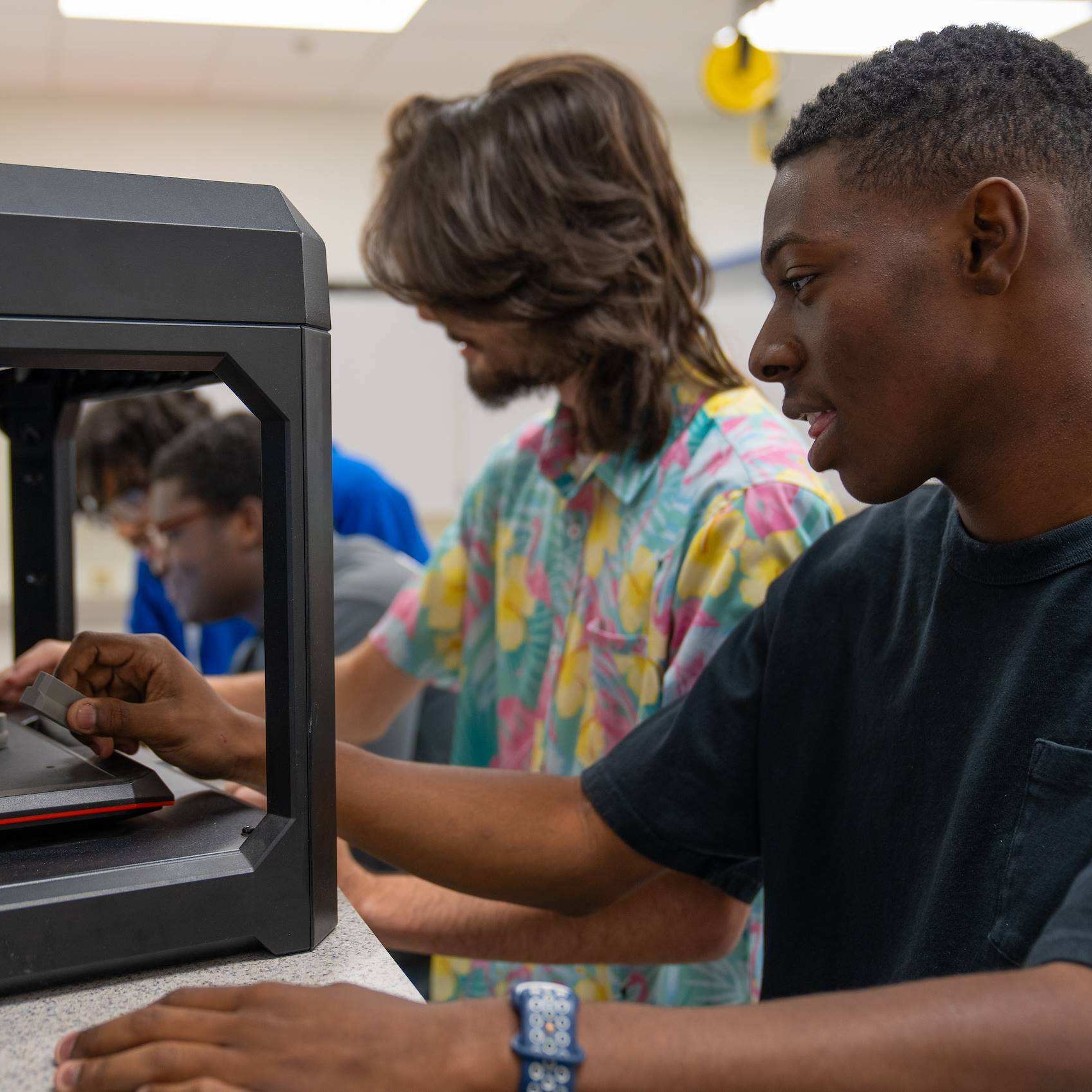 Three people examine a black 3D printer. One person operates the machine while another looks on.