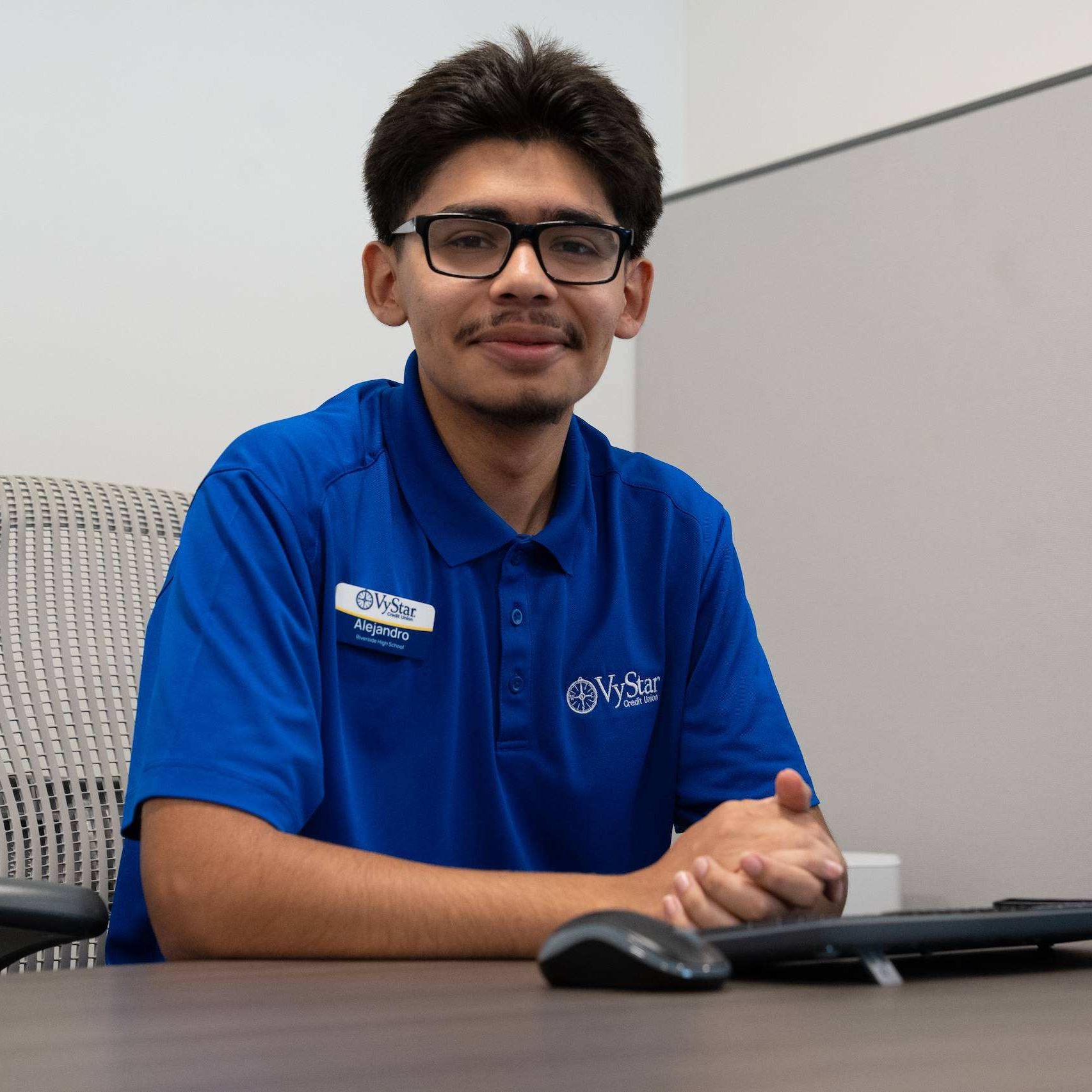 Man in a blue shirt sits at a desk with a computer, smiling. Behind him is a gray wall.