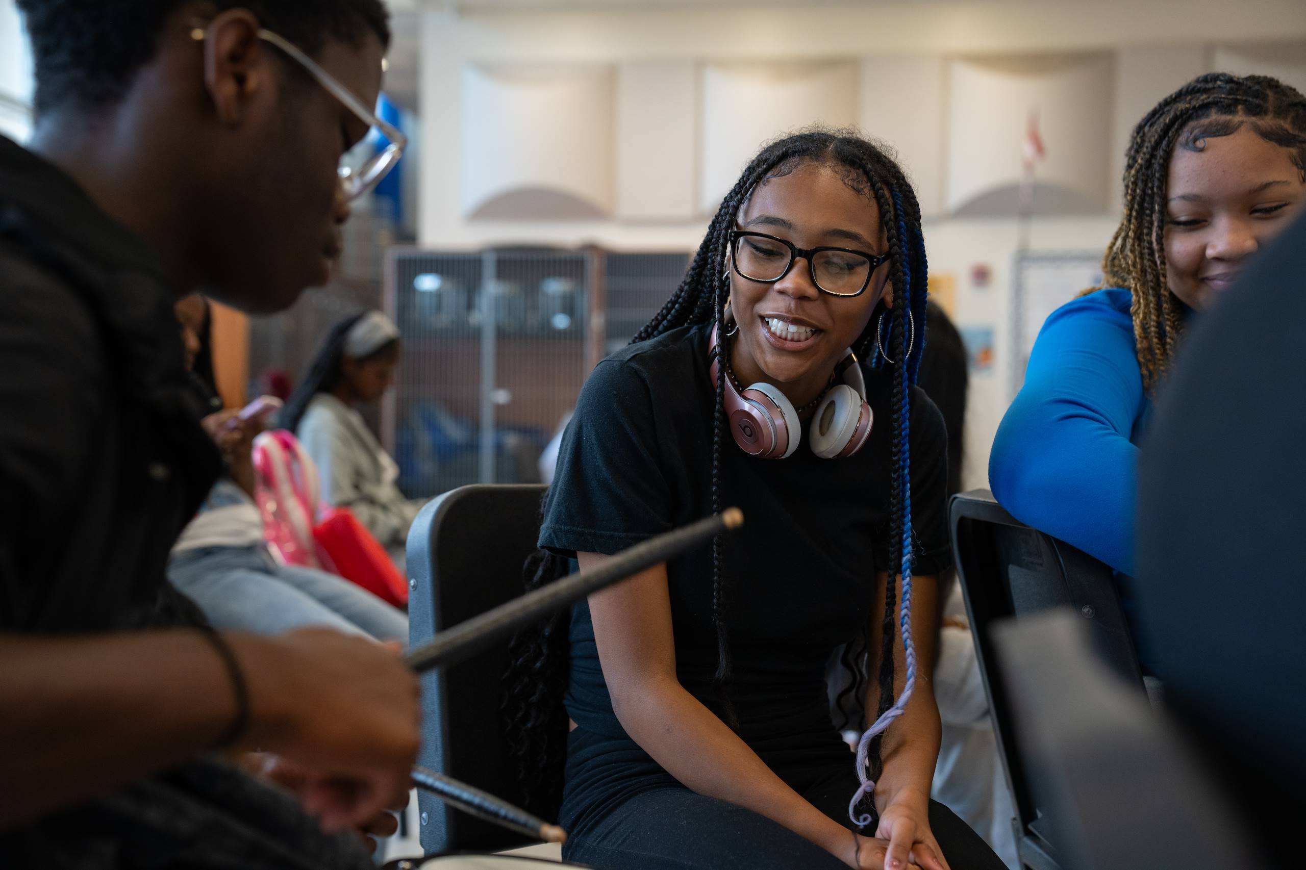 Two individuals, one wearing glasses, seated. One holding a drumstick, possibly engaging in conversation. Background: a room with other people.