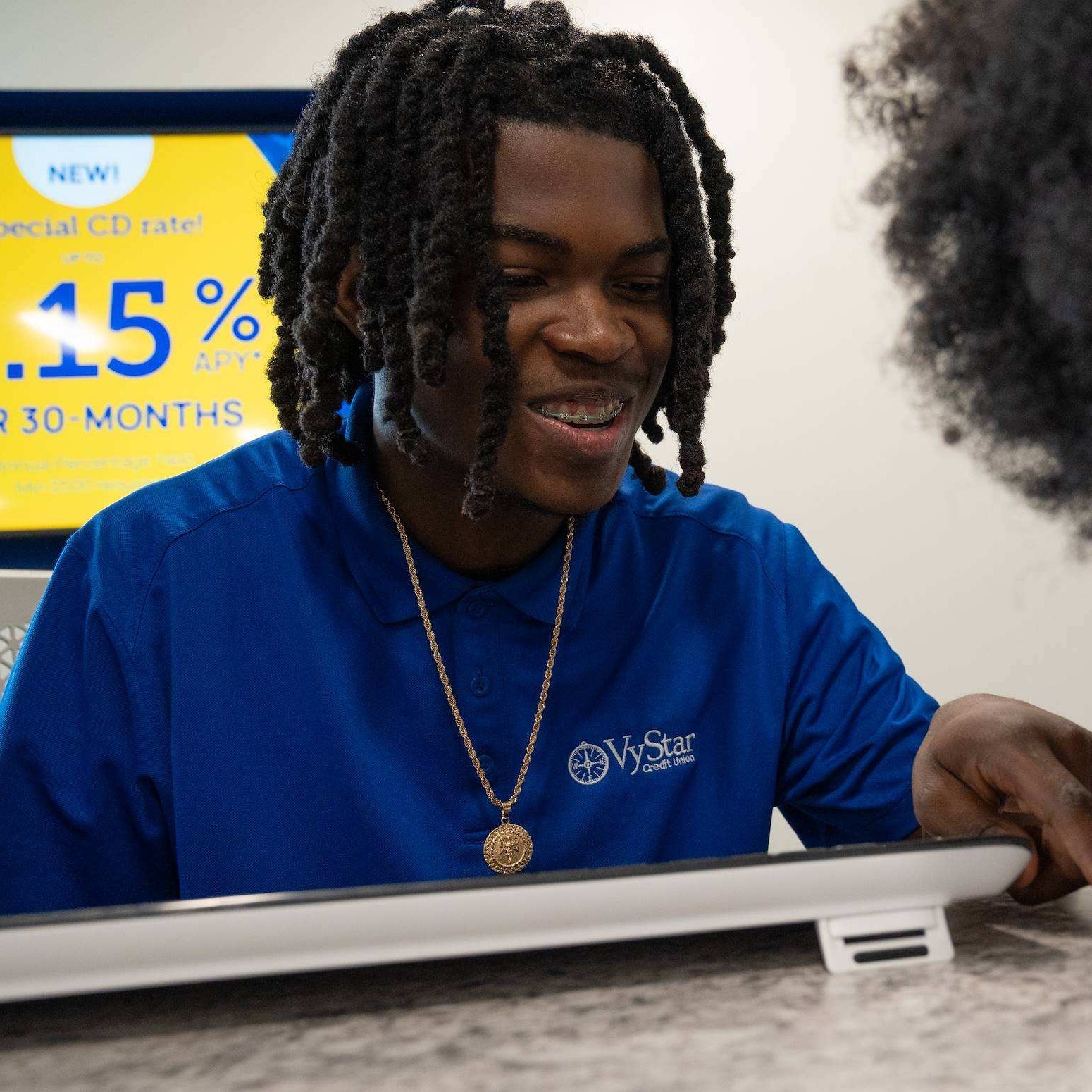 Two individuals focus on a paper, one with a blue shirt, a necklace, and a tablet, the other with glasses and a pen.