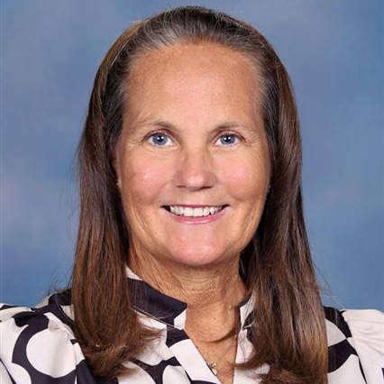A woman with brown hair wearing a black and white blouse. She has a warm smile and is standing against a blue background.