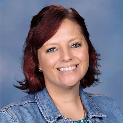 Woman with short red hair, wearing a denim jacket and floral top, smiling against a plain blue background.