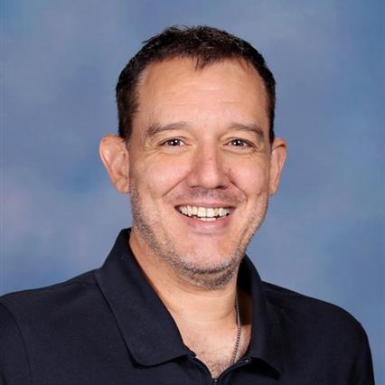 A man wearing a black polo shirt stands against a blue sky background, smiling and looking directly at the camera.
