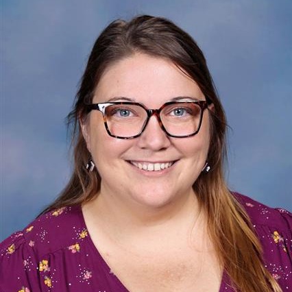 A woman with brown hair and glasses smiles against a light blue background. She wears a purple floral top.