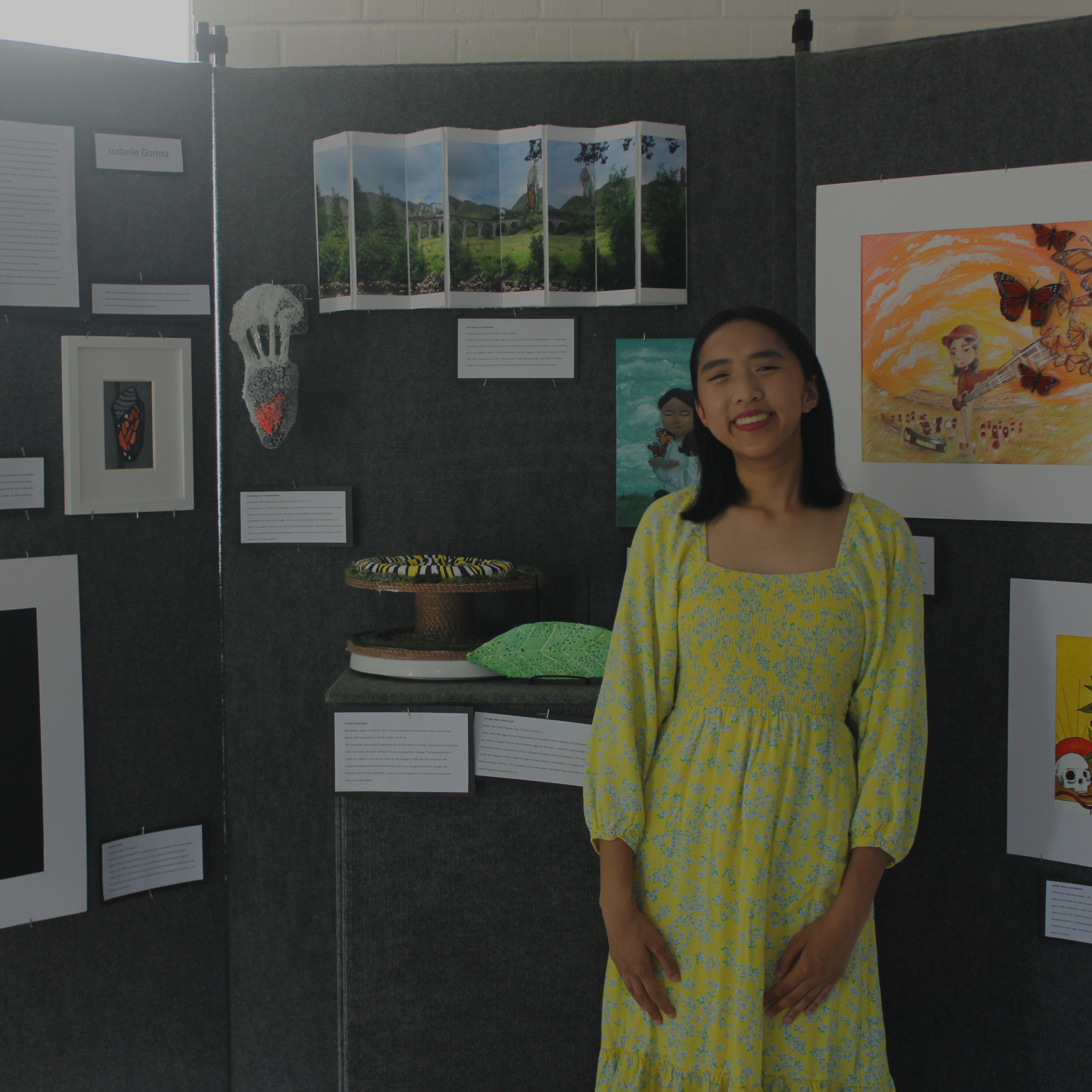A woman in a yellow dress stands before a display of framed artwork on a gray wall.