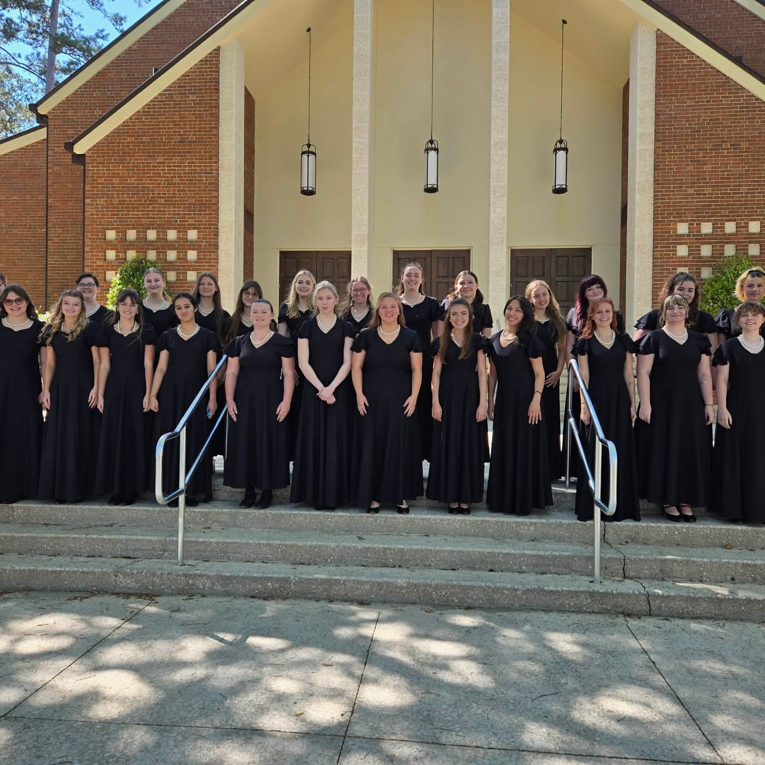 A choir in black dresses, lined up in rows, stands on the steps of a brick building.