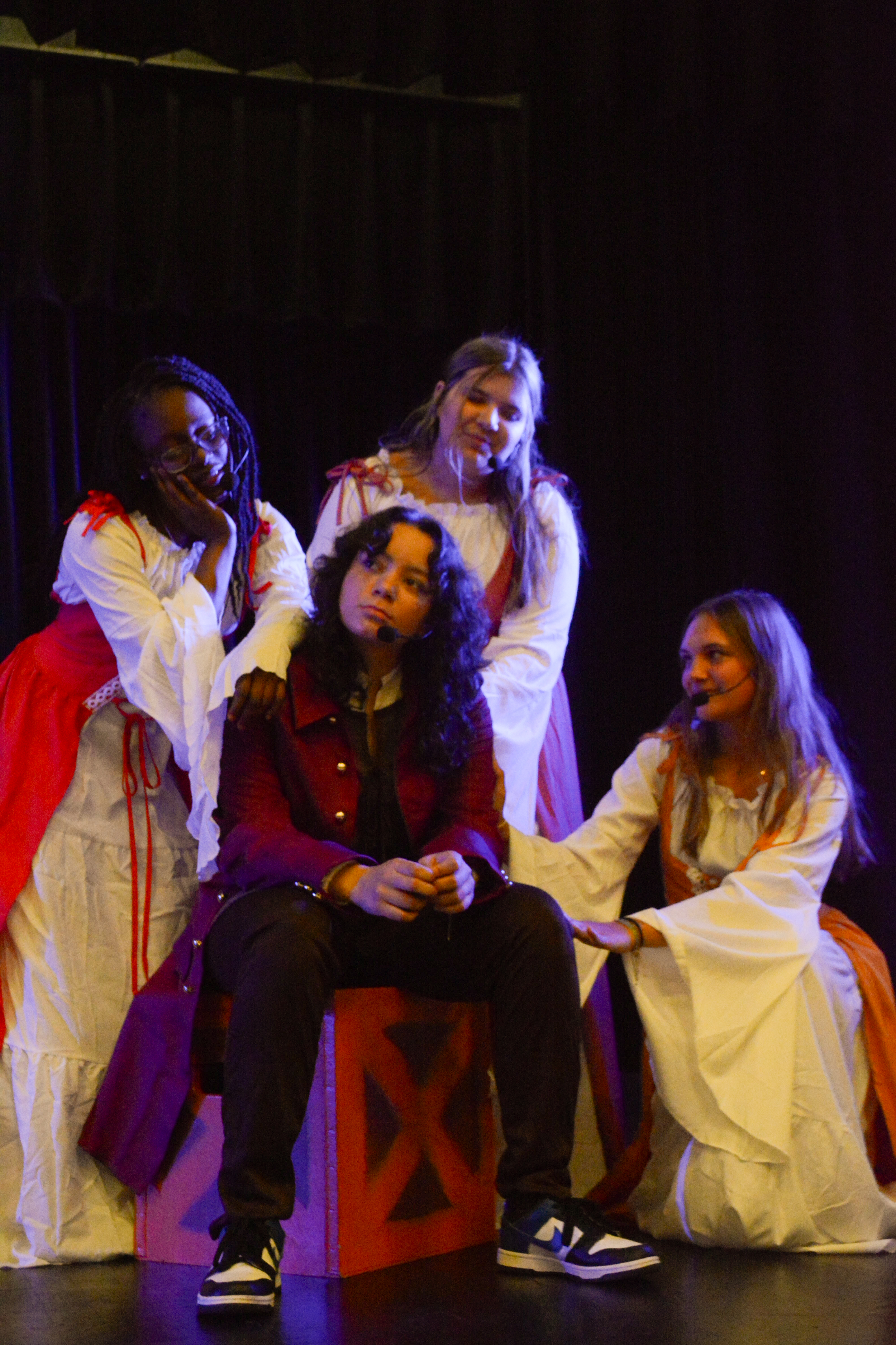 Four people on stage. One man in red with long dark hair. Three women with long hair in white and red.