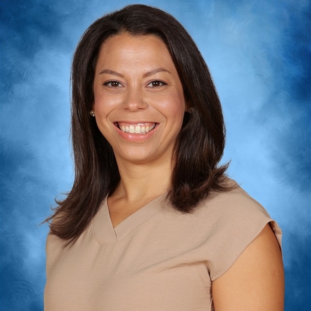 A smiling woman with brown hair and a tan top poses against a solid blue background.
