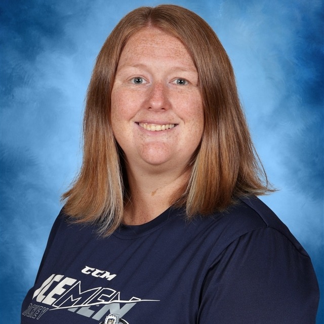 An adult woman with blonde hair smiles, standing in front of a blue background. She wears a navy blue shirt with white text.
