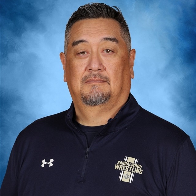 Man in navy blue jacket with white "Under Armour" logo and "Sandelwood Wrestling" patch. He looks straight ahead.
