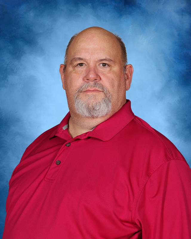Man with a beard, short hair, and mustache. Wearing a red polo shirt. Standing in front of a blue background.