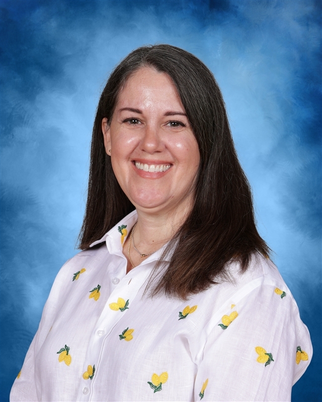 A woman with long dark hair smiles at the camera. She wears a white floral button-up shirt.