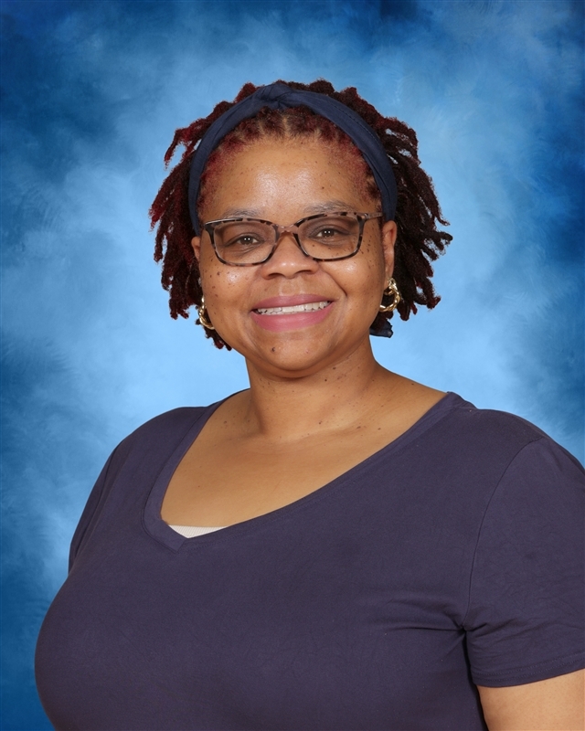 Person with curly hair, wearing a headband, glasses, and a purple shirt. Standing against a blue background.