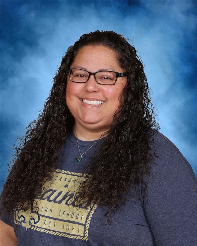 A woman with curly dark hair and glasses smiles at the camera. She wears a gray shirt with a design and text.