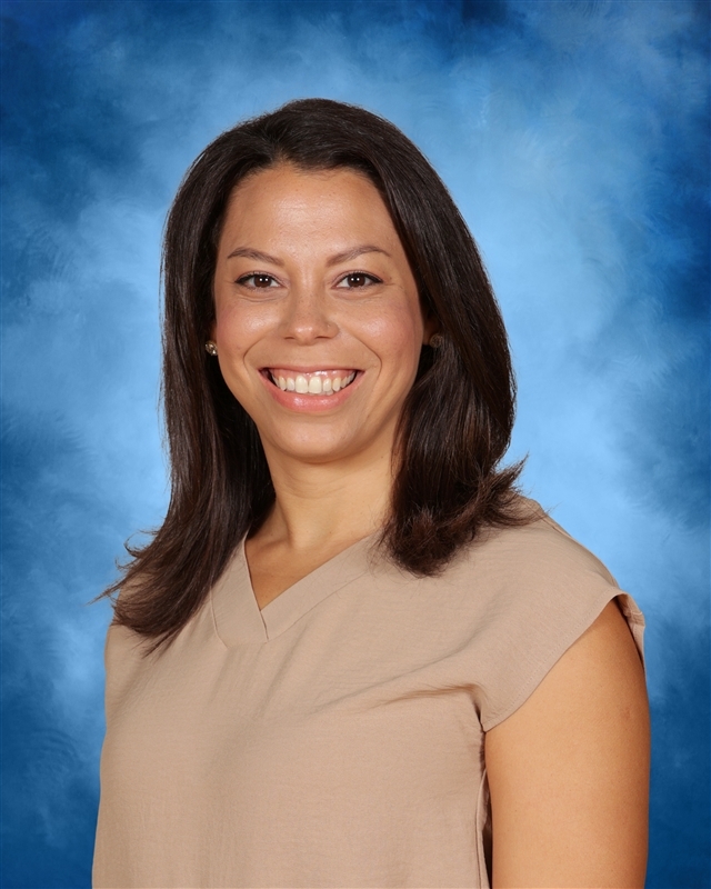 Woman smiling, standing in front of a blue background. She wears a tan V-neck top and has long, dark hair.