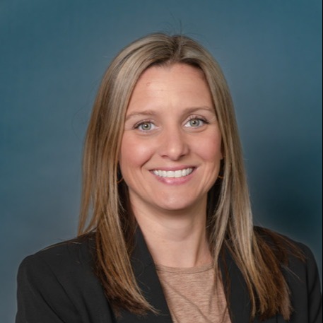 A woman with long blonde hair smiles, wearing a black blazer over a brown top, against a blue background.