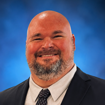 A man with a beard, bald, wearing a dark suit, light shirt, and patterned tie stands smiling against a blue background.