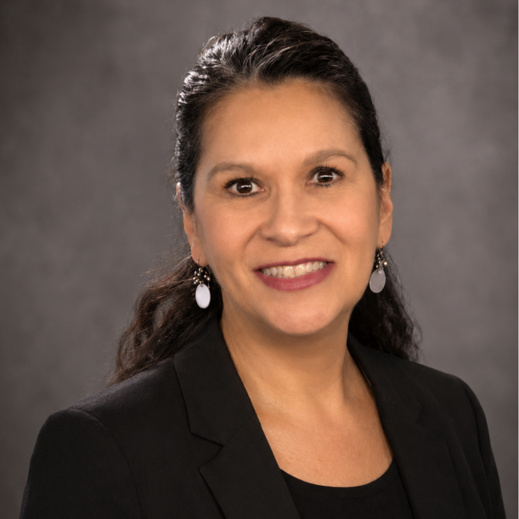 A woman smiles, standing against a gray background. She wears a black blazer over a black top and has earrings.