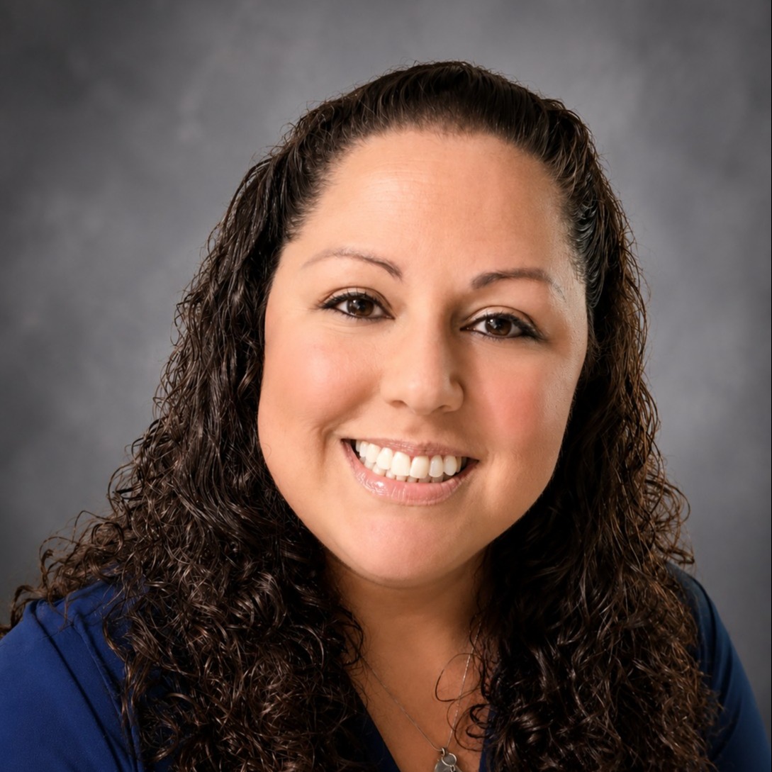 Woman with curly dark hair wearing a blue shirt and necklace, smiling with teeth visible.