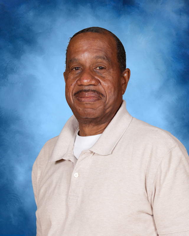 A man with a smile and short hair, wearing a light tan polo, stands against a blue background.