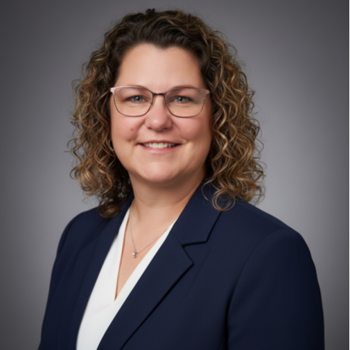 A woman with curly hair, wearing glasses, a white shirt, and a navy blue blazer. She smiles for the camera.