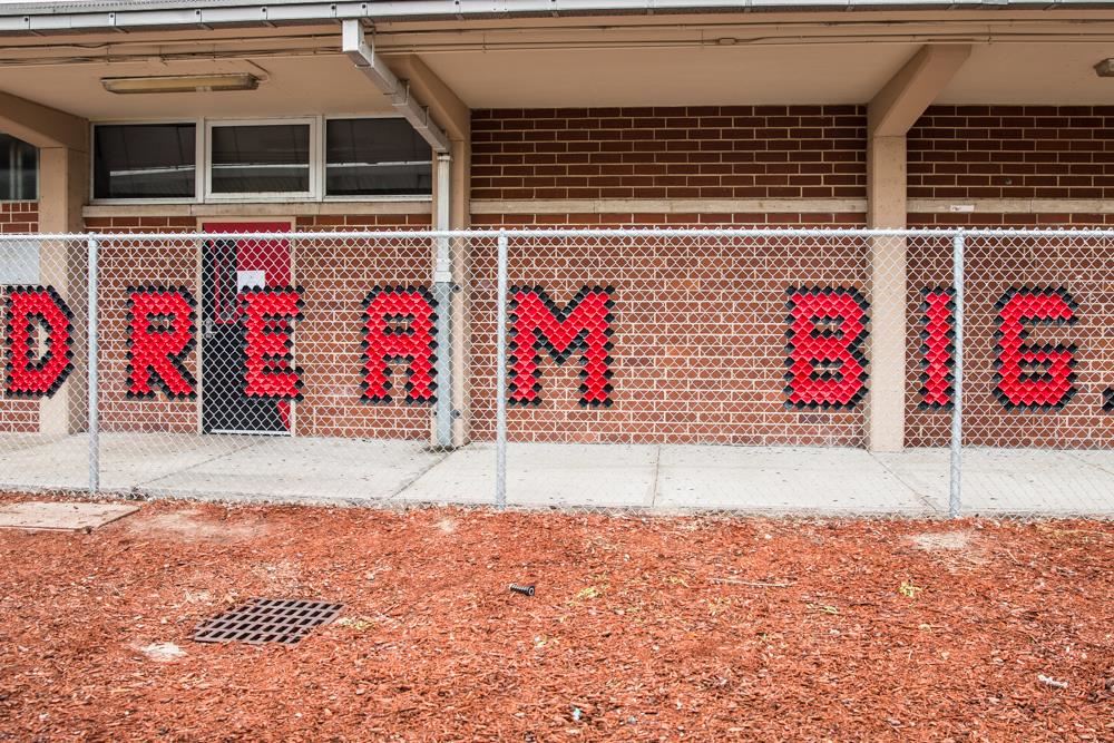 A brick building with a chain-link fence displays the words "DREAM BIG" in red letters on the wall.