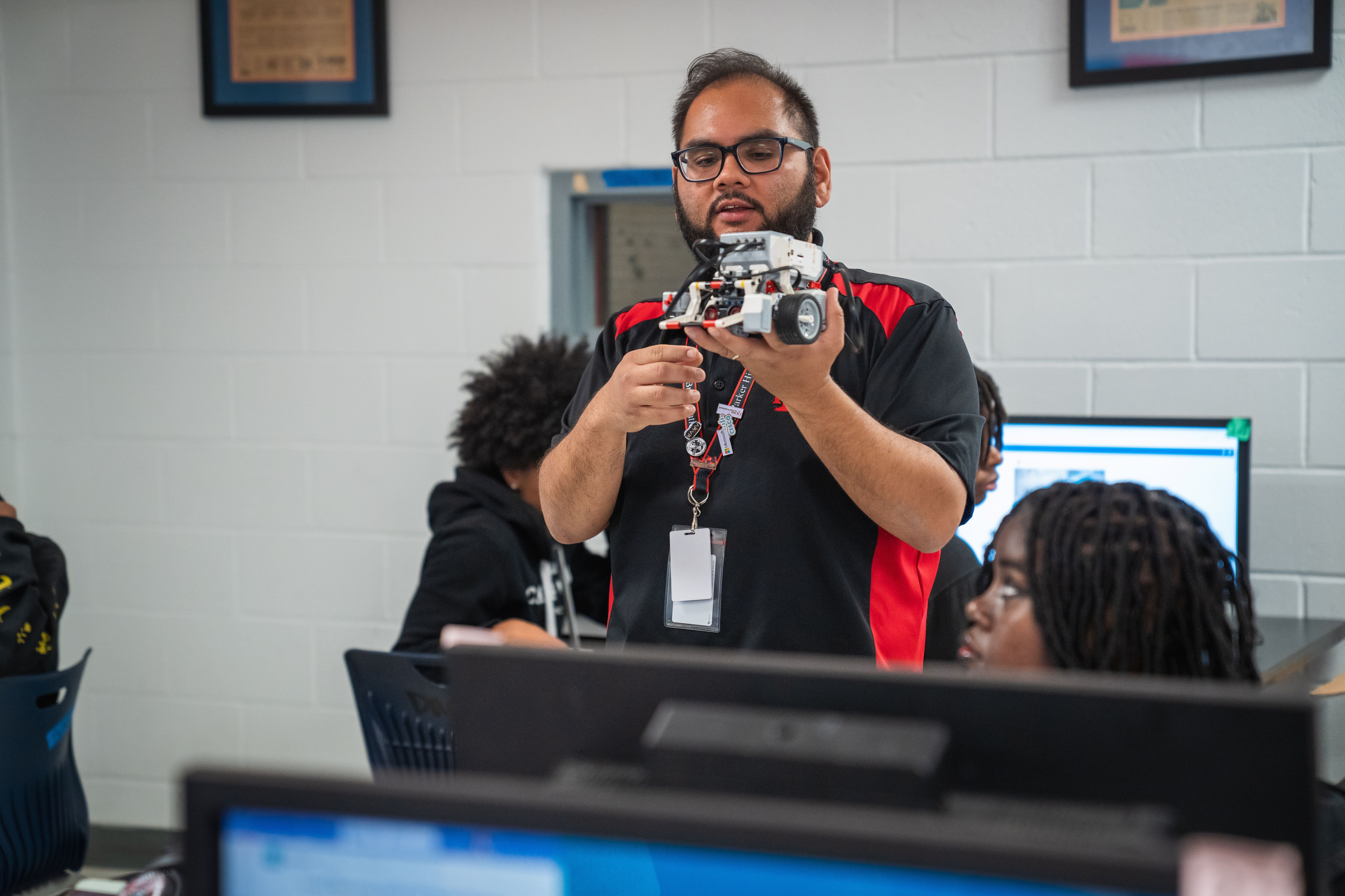 Man stands with camera in a classroom, holding it in front of a monitor. Students sit behind him, focusing on screens.