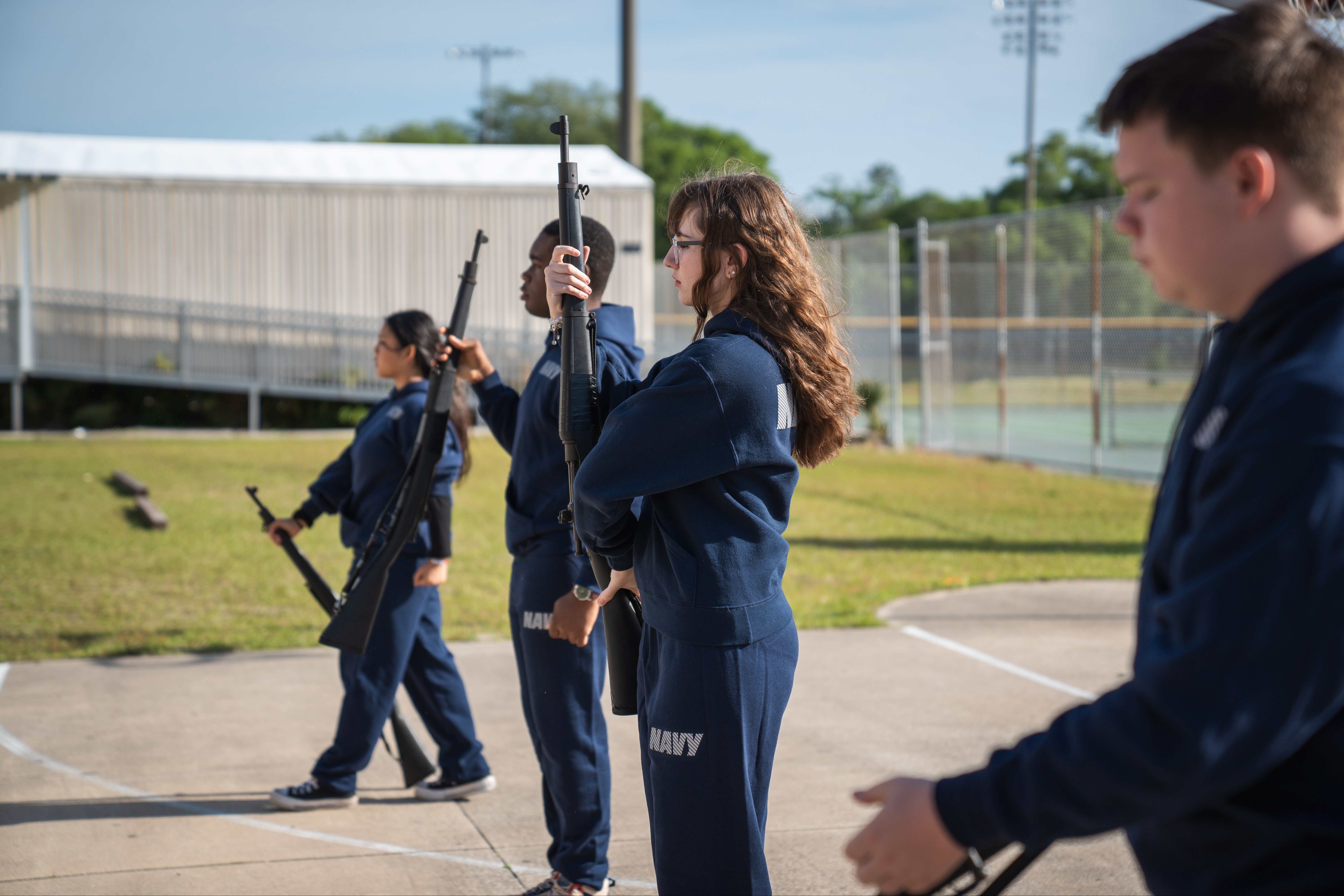 Four people in navy blue uniforms stand in a line, each holding a rifle, outside a building.