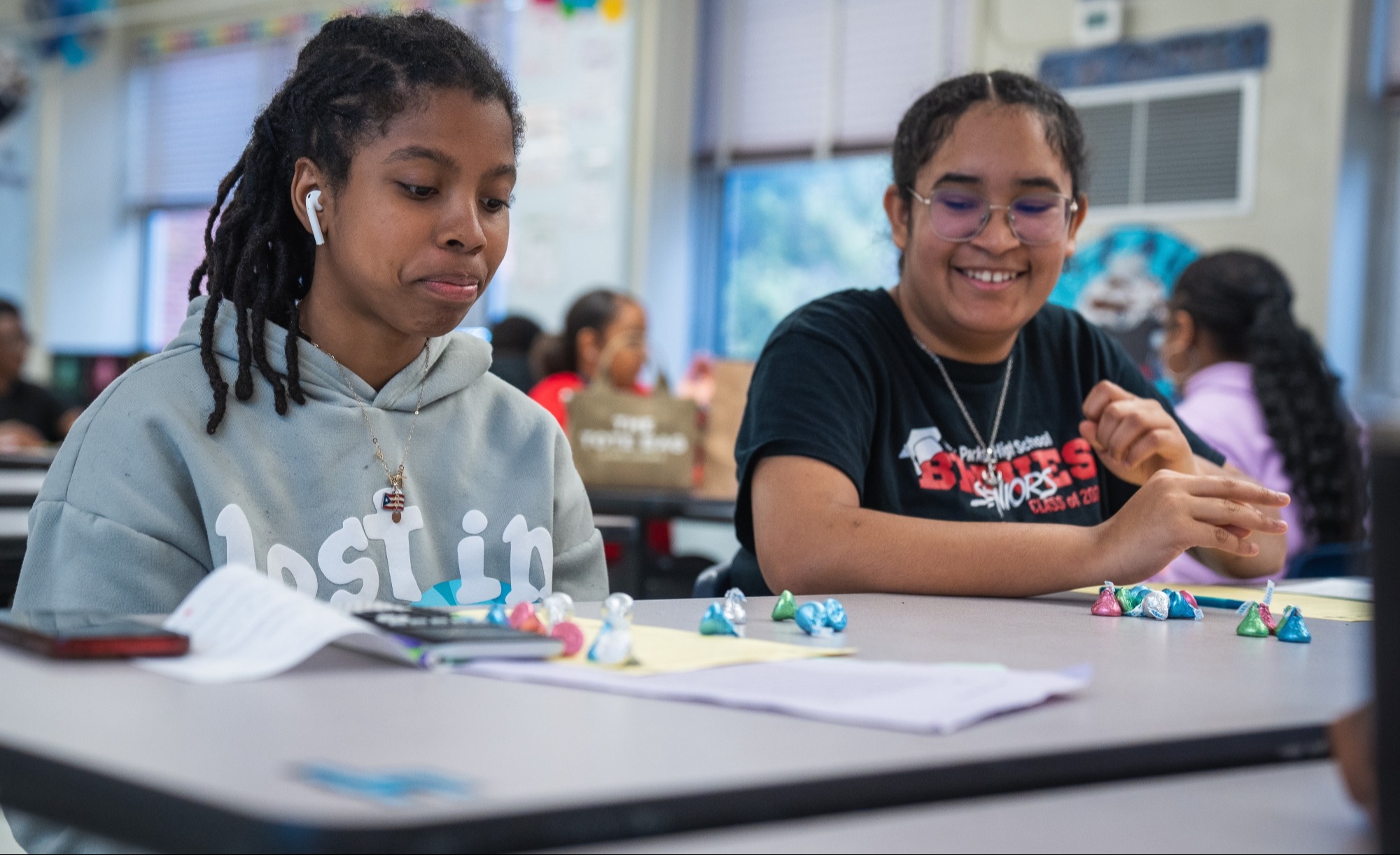 Two young girls in a classroom, one focused on her work, the other smiling, with dice and papers on the table.