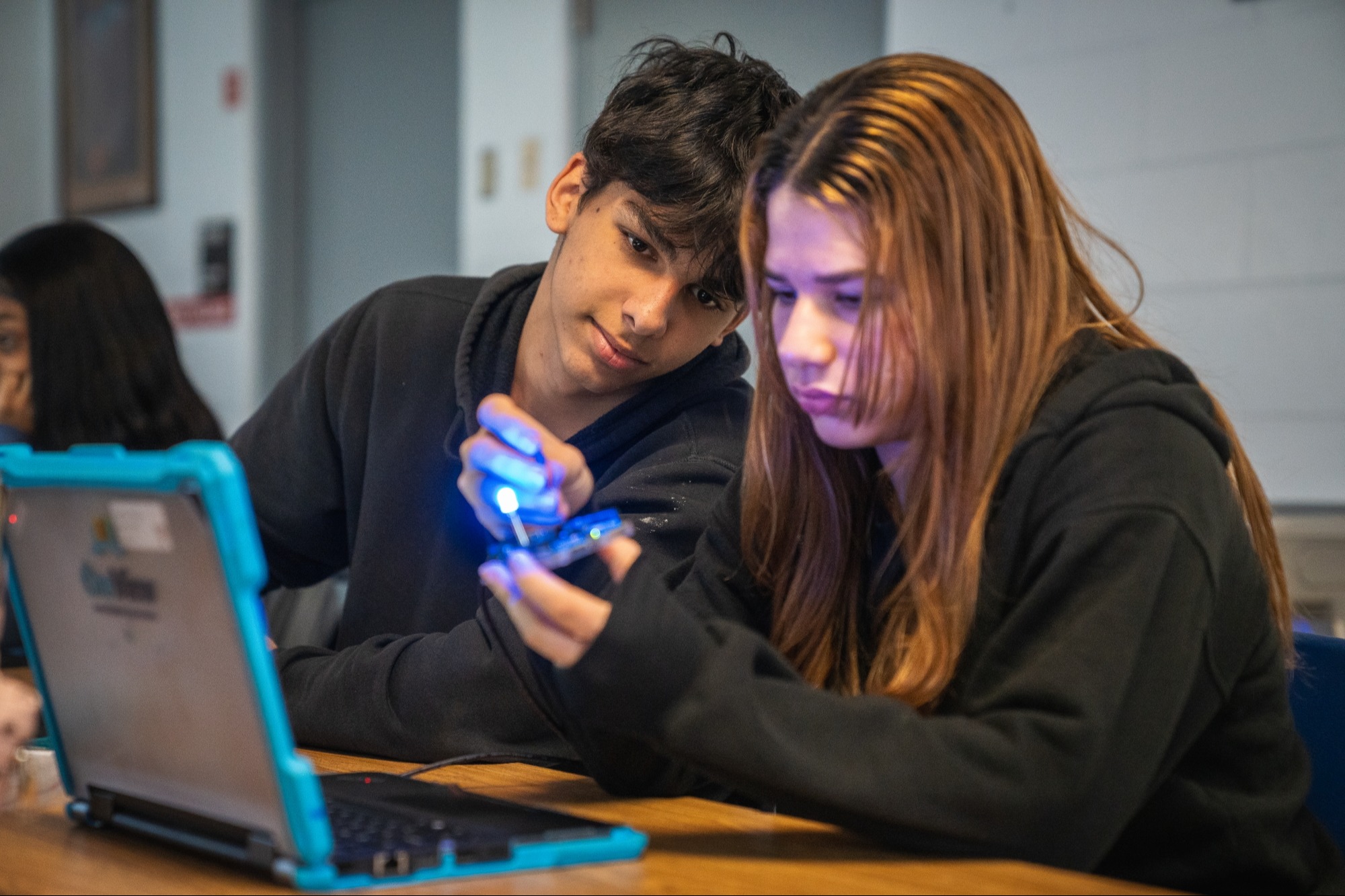 Two individuals focus on a laptop with a blue light; one holds a small device, both wearing dark hoodies.