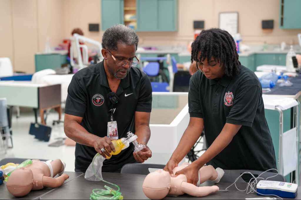 Teacher helping a student learn CPR procedures on a pretend baby
