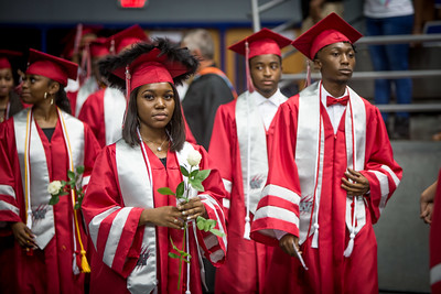 Students walking in graduation caps and gowns, girl on left holding a white rose