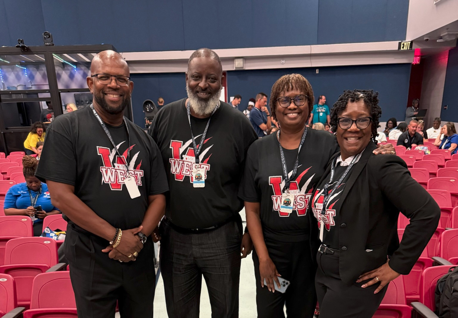 Four people stand in a row in a stadium with red chairs, wearing black t-shirts with "WEST" on them.
