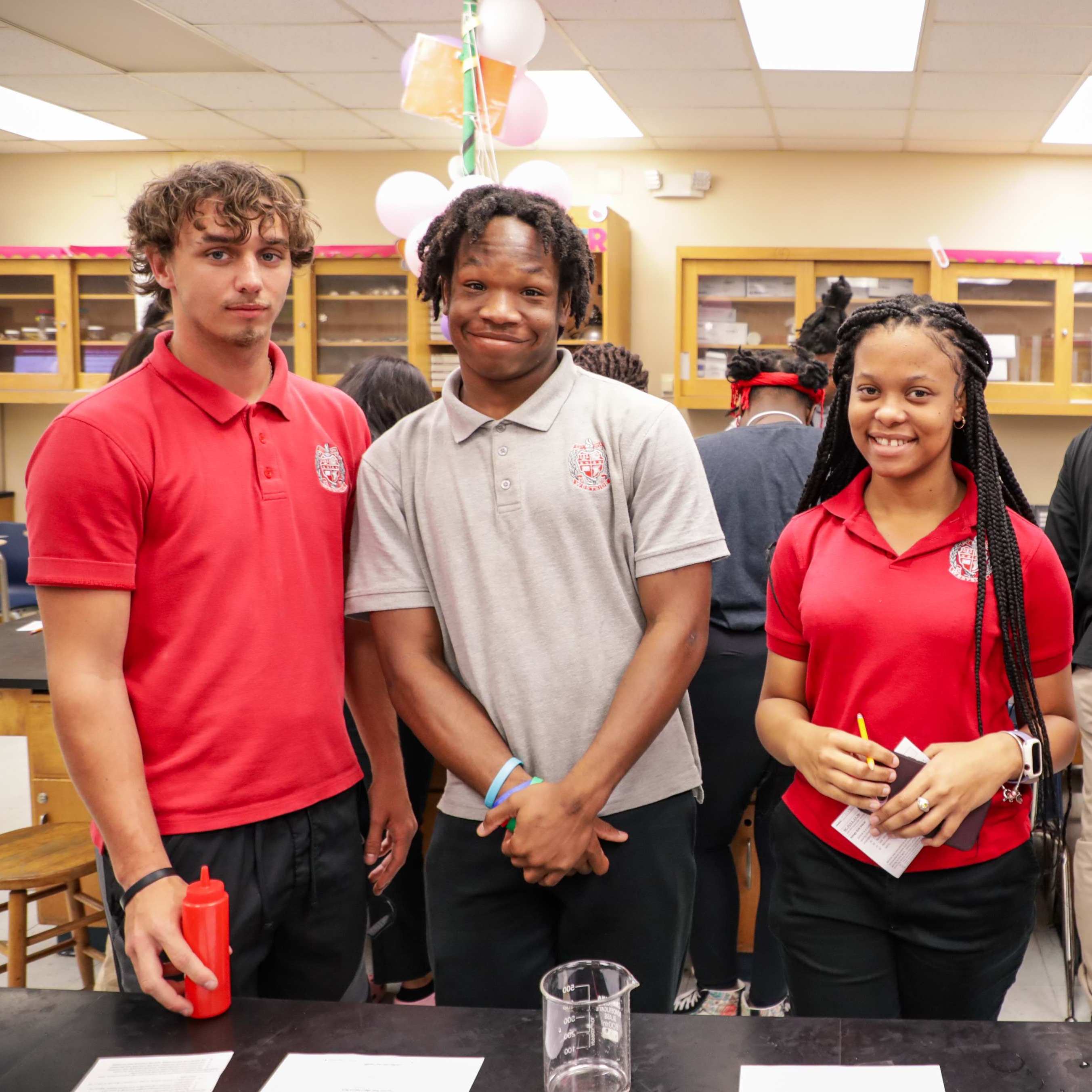 A group of four young adults, wearing red shirts, standing in a classroom and smiling at the camera.