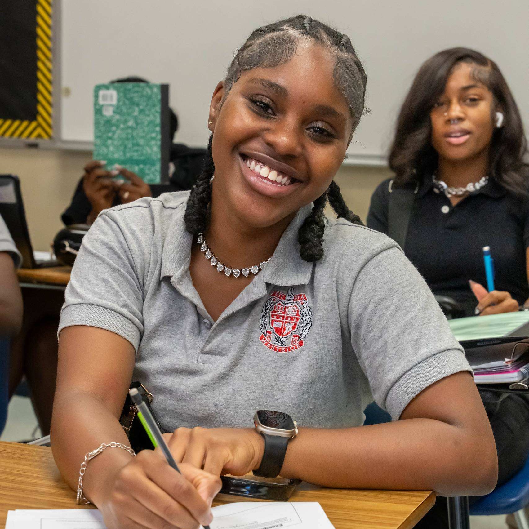 A girl in a grey polo shirt sits at a desk, smiling, and writes in a notebook.