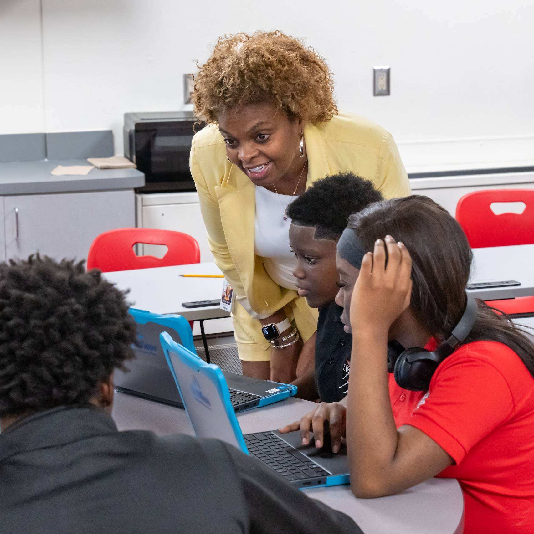 Five students sit at a table with laptops. A woman in a yellow sweater stands behind them, smiling.