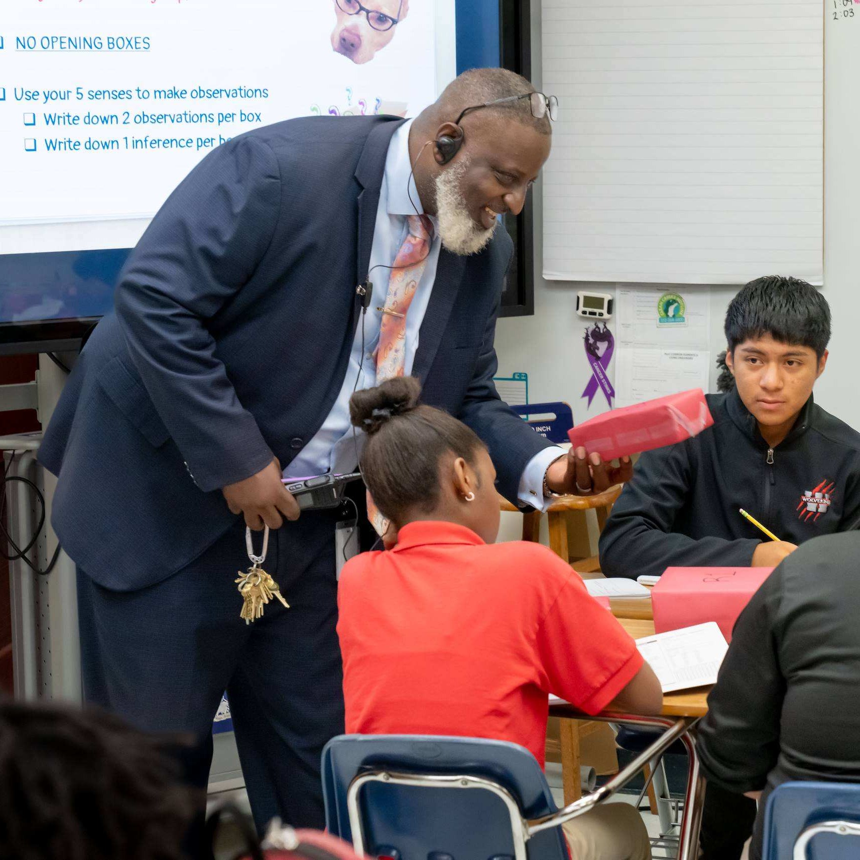 A man stands at a desk and helps a group of children seated around it, in a classroom.