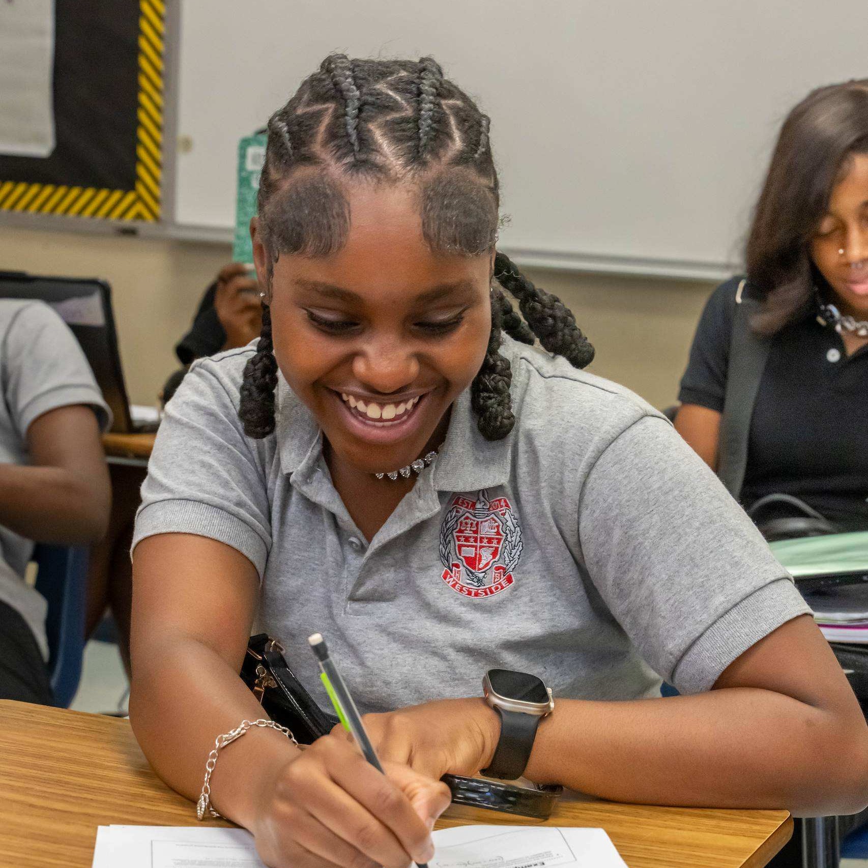 A girl sits at a desk, smiling, and writing on paper with a pen in a classroom.