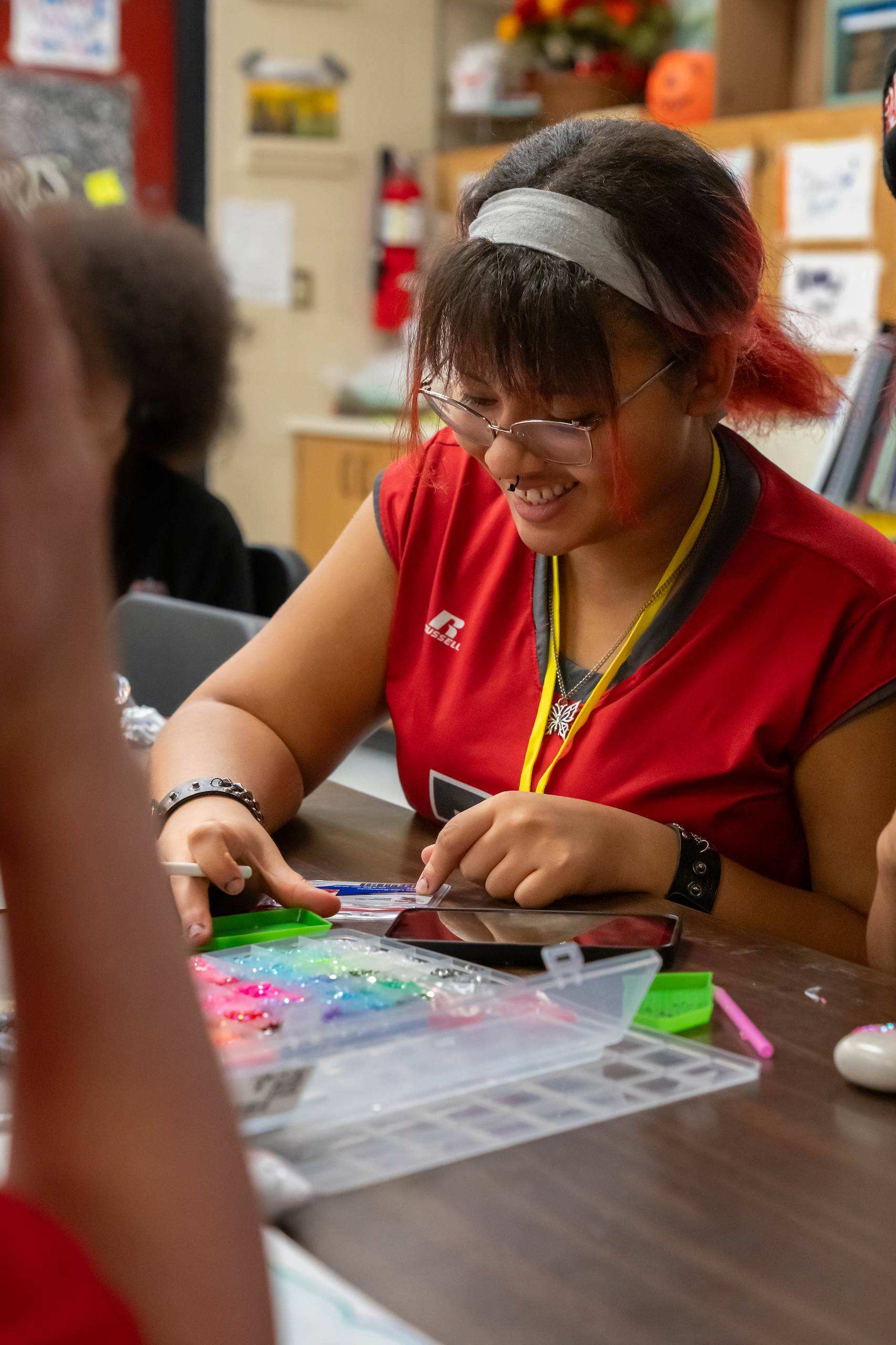 A woman in a red shirt and glasses sits at a table, smiling while working on a craft project.