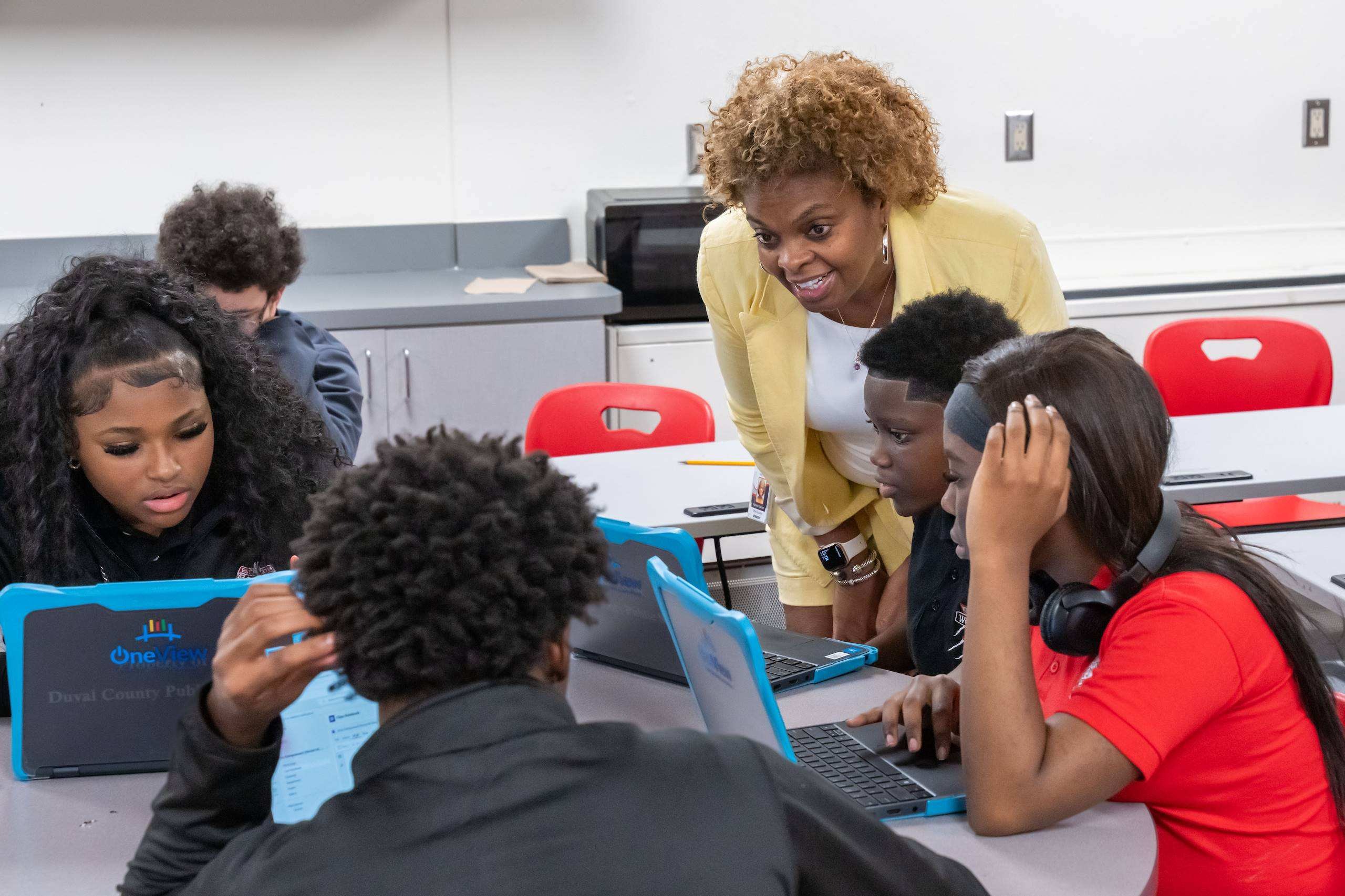 Group of students in a classroom. One adult in yellow is teaching them while they use laptops.