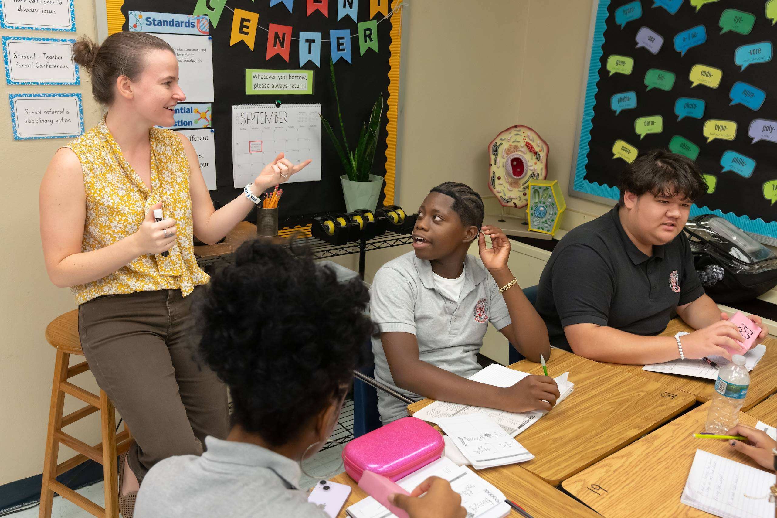 Teacher speaking to students in a classroom with a bulletin board in the background.