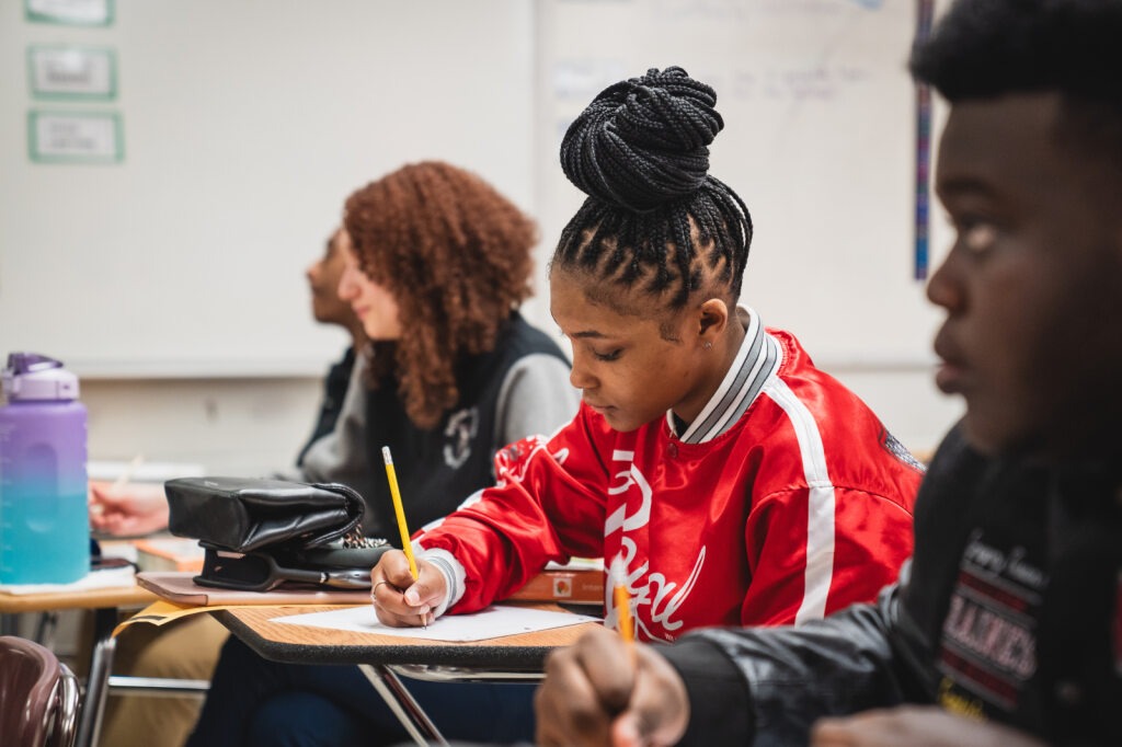 A young person in a red jacket writes on paper in a classroom. A student sits behind them.