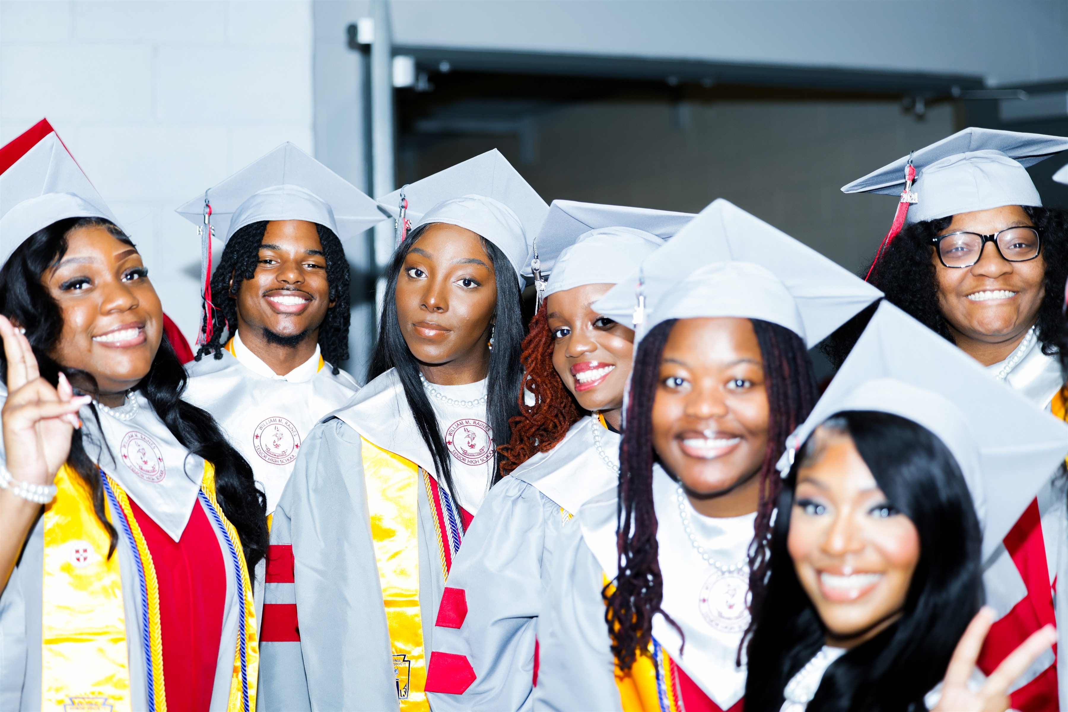 A group of individuals in academic regalia, including gowns and caps, stand together smiling. Some make hand gestures.