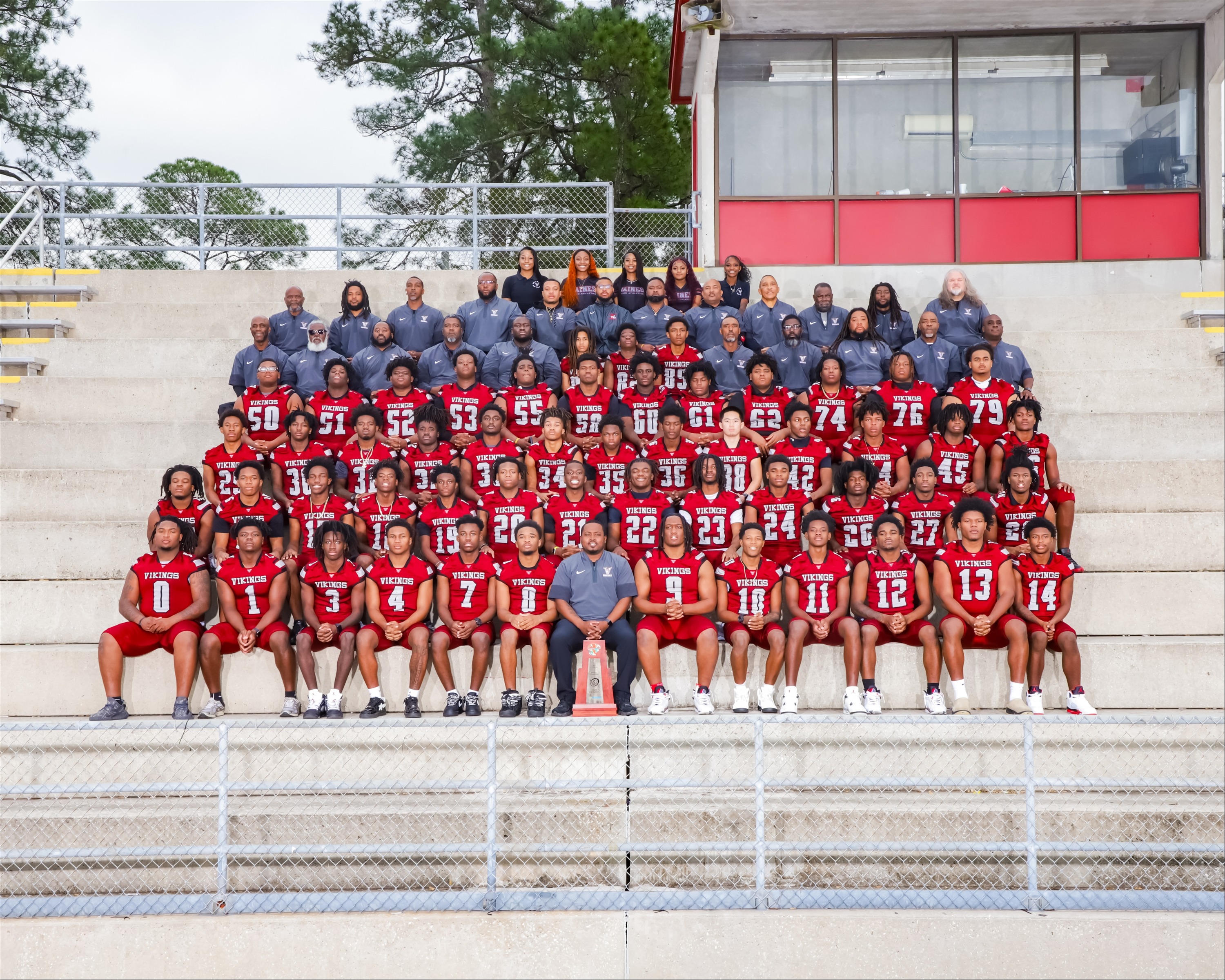 A team of football players in red jerseys pose on a stadium bench with a red background.