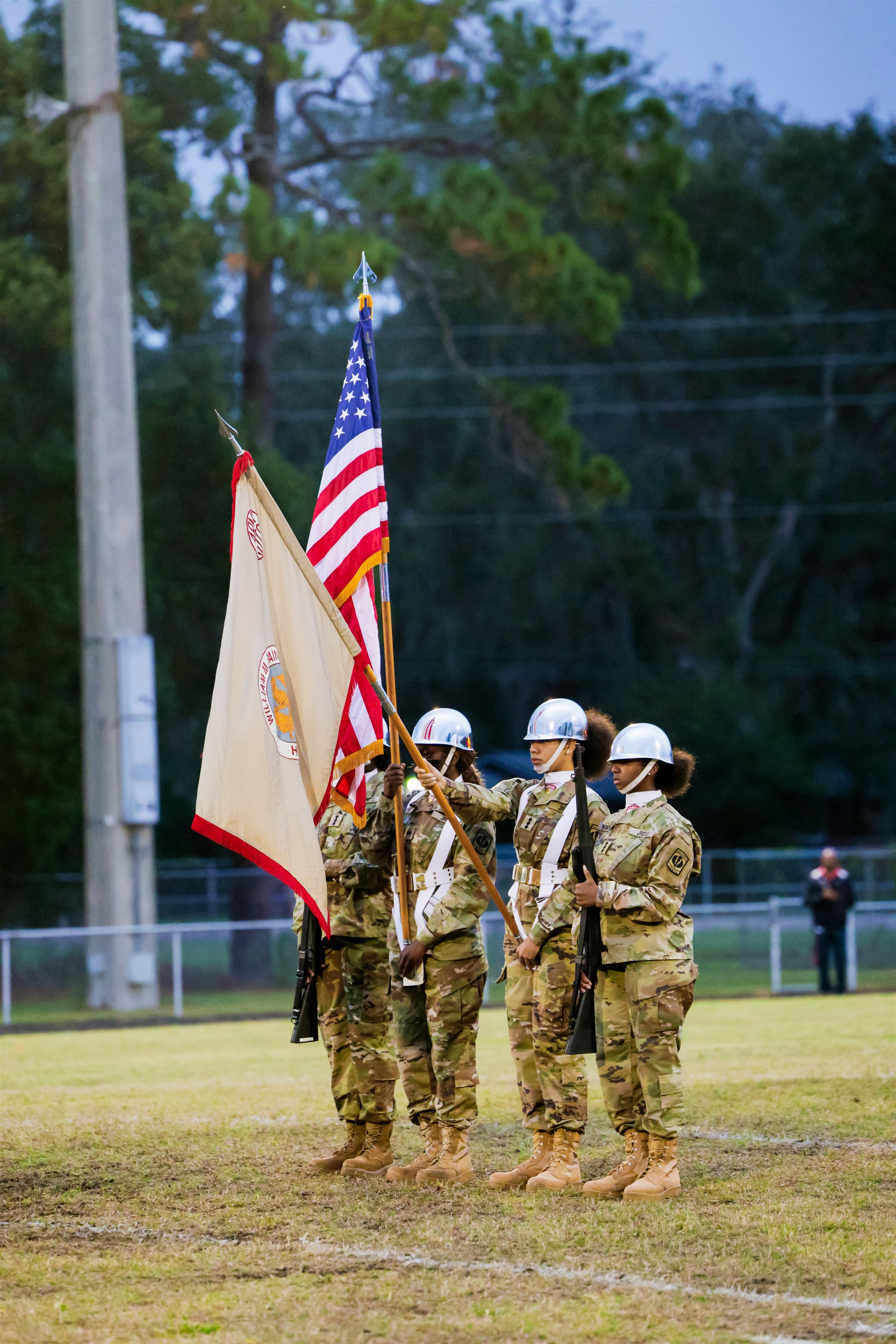Five military personnel hold flags, including a US flag, on a grassy field. A metal pole and trees are in the background.