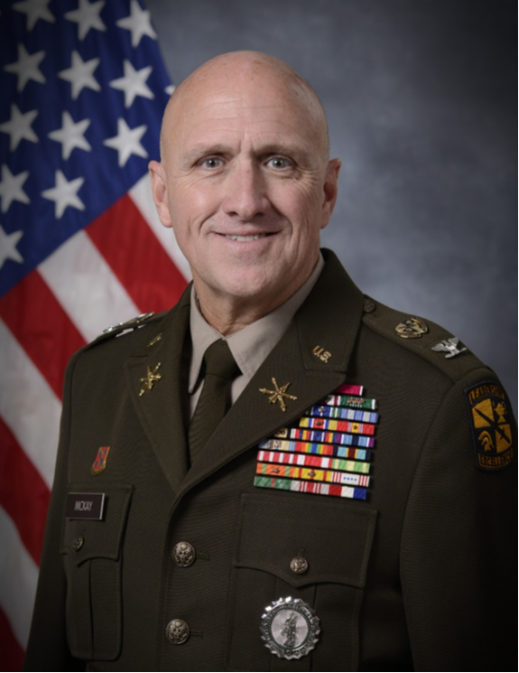 Man in a military uniform with medals stands in front of an American flag. He has a friendly expression.