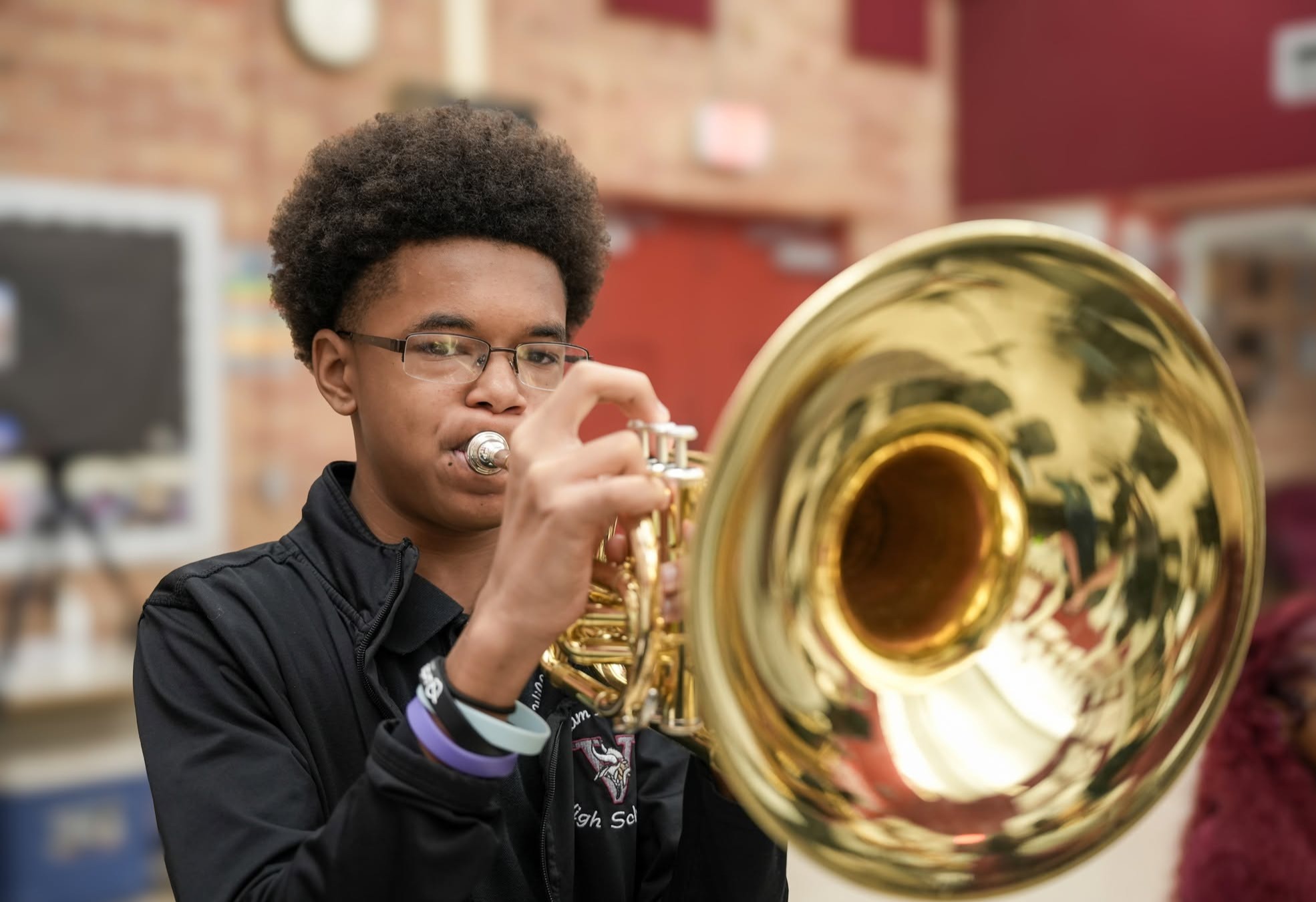 A young person plays a golden trumpet while standing indoors. A second person in a red shirt is partially visible.