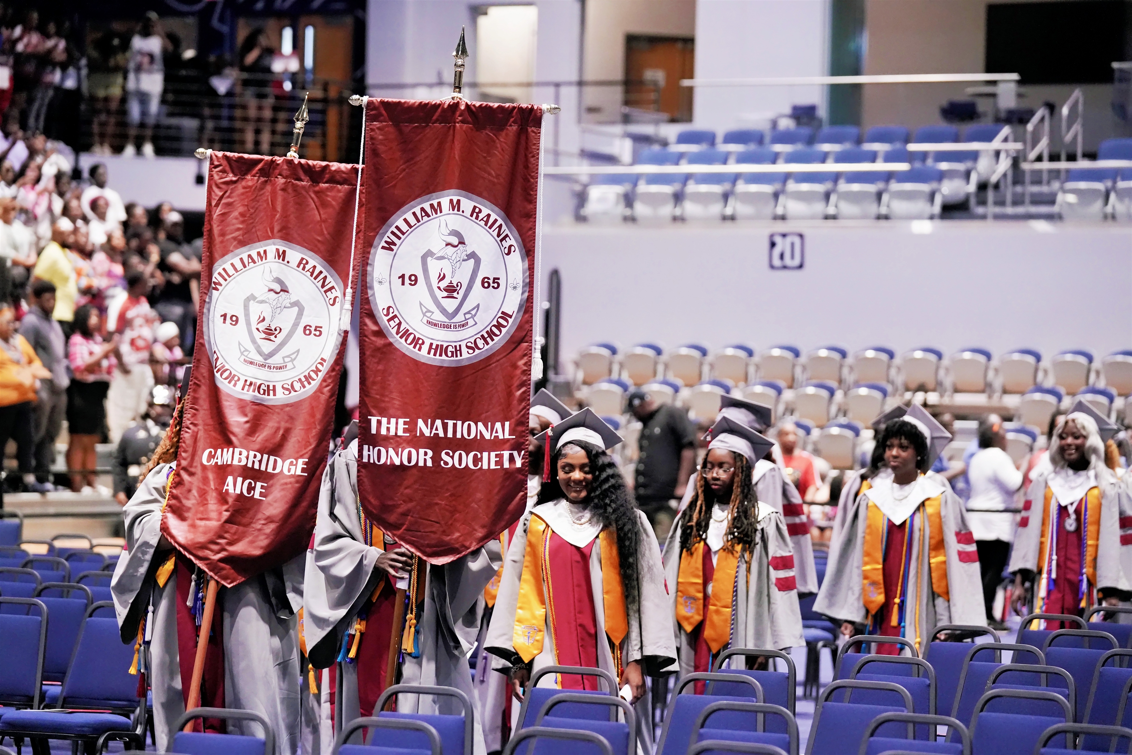 Graduates in caps and gowns walk in a stadium. Two hold red banners with text. Audience sits on blue chairs.