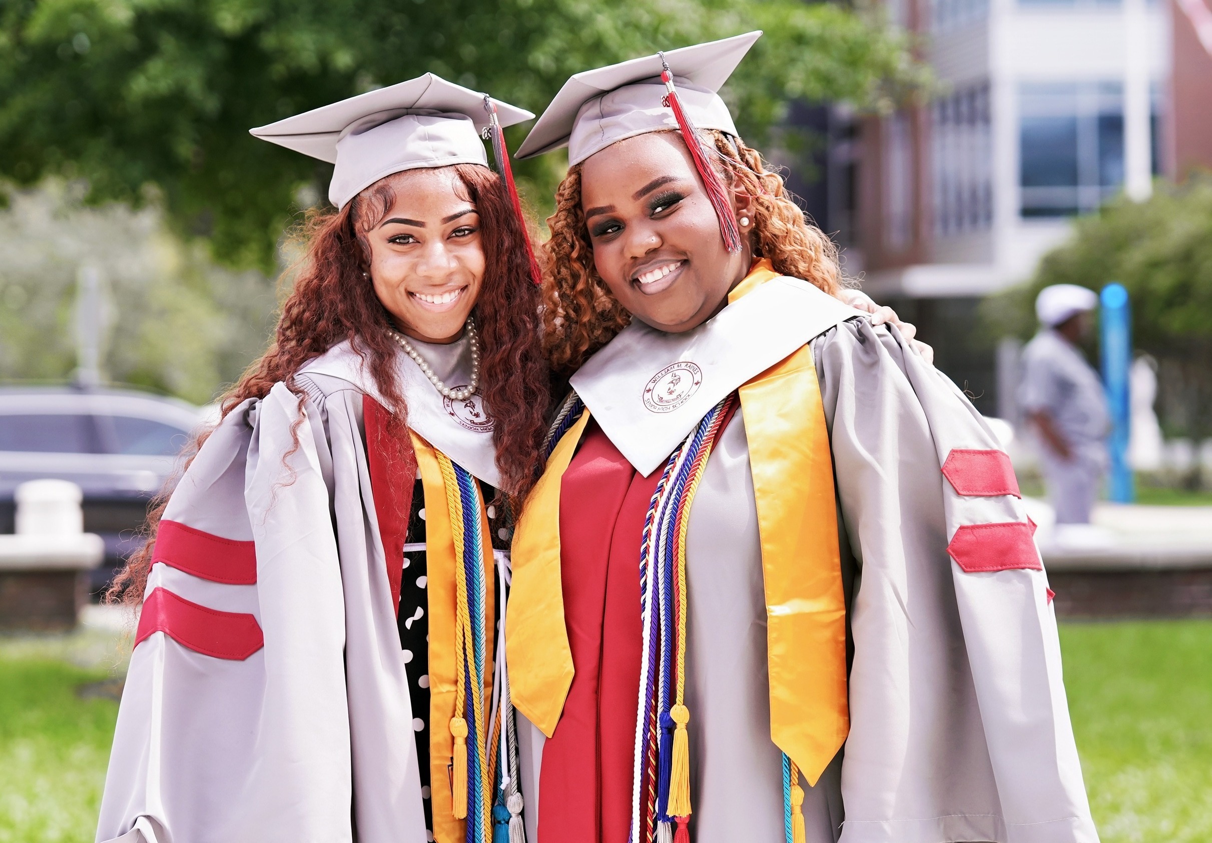Two women in graduation gowns stand on a grassy lawn in front of a building. One wears a gray robe.